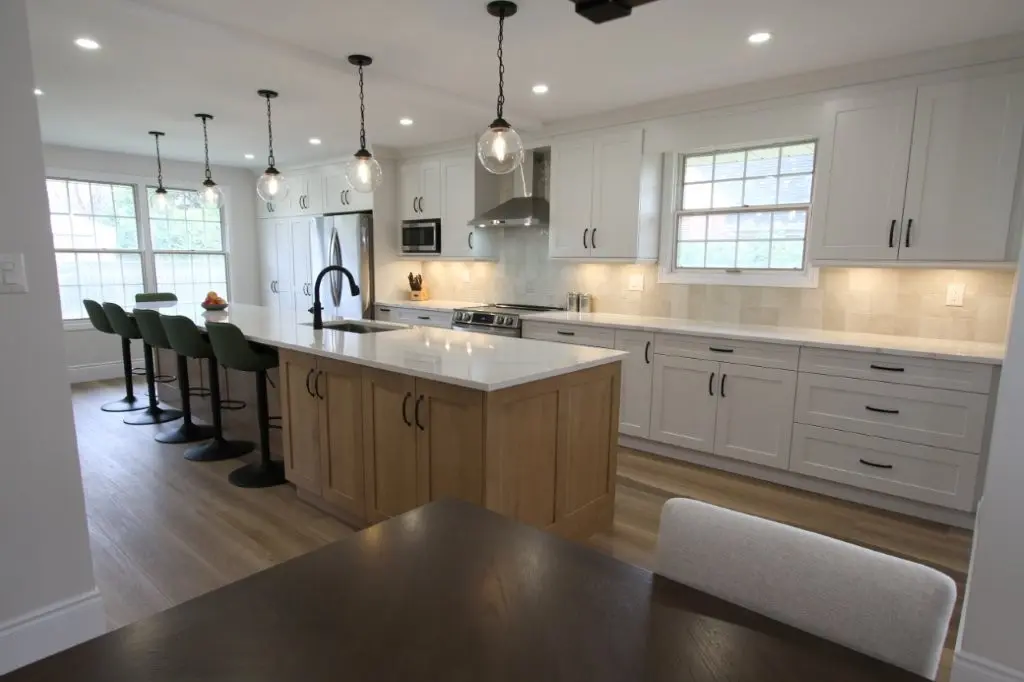 A modern kitchen with white cabinets, stainless steel appliances, and a large island with barstools. The island features a wooden base and a white countertop with a built-in sink and faucet. Pendant lights hang above the island, and there is a window in the background letting in natural light.