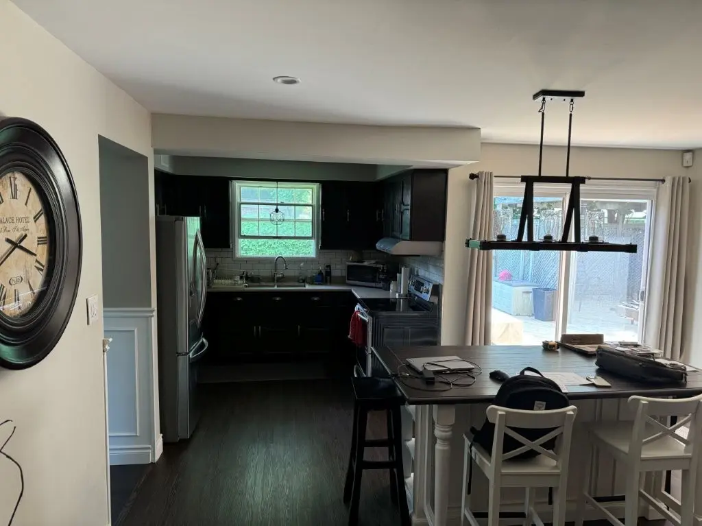 A before renovation kitchen with dark cabinets, a window above the sink, stainless steel appliances, and an island with high chairs. A large wall clock is mounted to the left, and there is a sliding glass door with curtains on the right leading to an outdoor area.