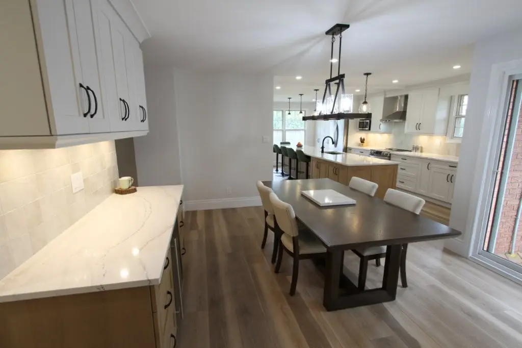 Modern kitchen and dining area with white cabinets, quartz countertops, a dark wooden dining table with chairs, and a large island with bar stools, all illuminated by prominent light fixtures.
