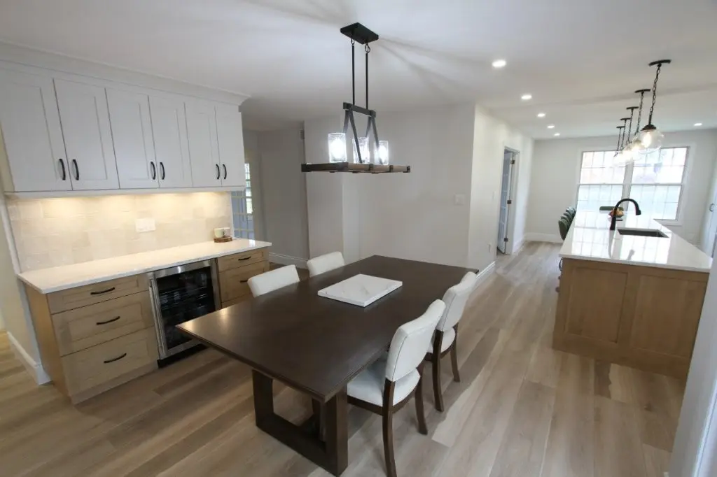 A modern kitchen with a wooden dining table and white chairs, under a rectangular chandelier. Light-colored hardwood floors, white cabinets, and a kitchen island with pendant lighting are also visible.