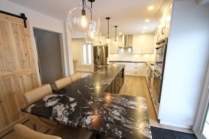 Modern kitchen with black granite countertops, light wood flooring, white cabinets, stainless steel appliances, tan leather dining chairs, hanging glass pendant lights, and a sliding wooden barn door.