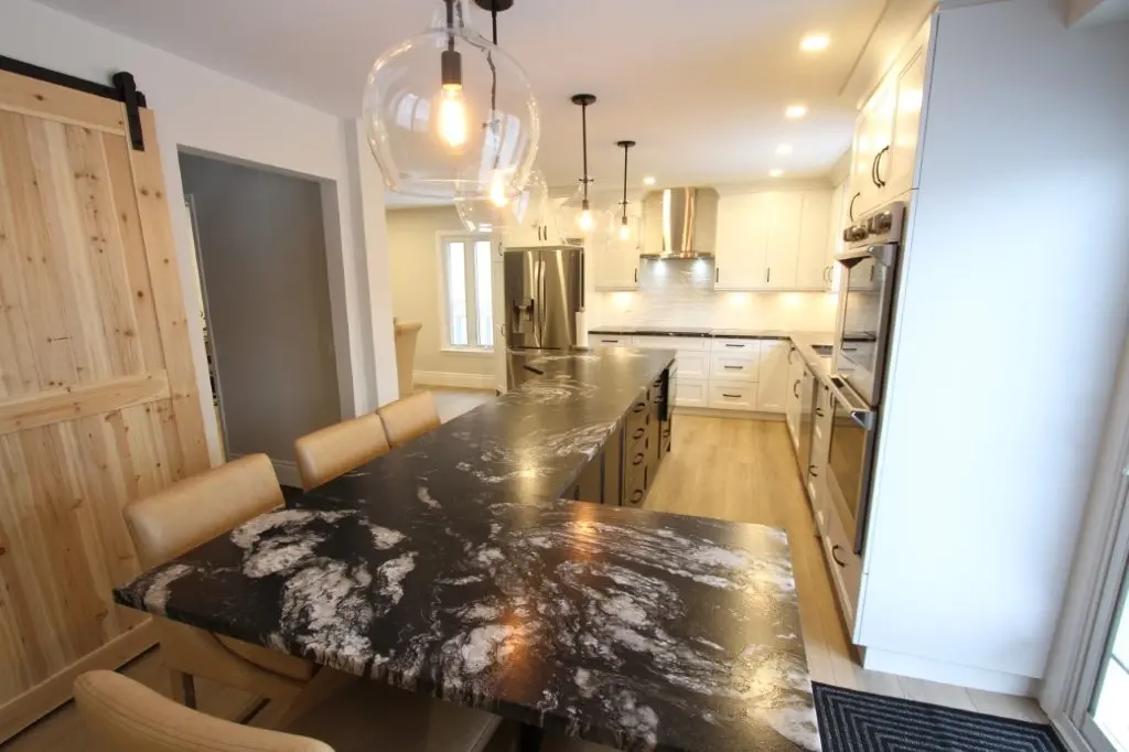 Modern kitchen with black granite countertops, light wood flooring, white cabinets, stainless steel appliances, tan leather dining chairs, hanging glass pendant lights, and a sliding wooden barn door.