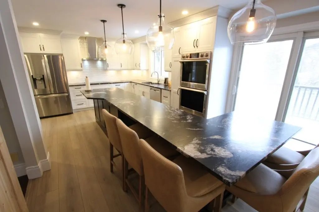 Modern kitchen with a long black and white granite island countertop surrounded by six beige upholstered chairs, white cabinetry, stainless steel appliances including a refrigerator and double oven, light wood flooring, three glass pendant lights, and a sliding glass door letting in natural light.