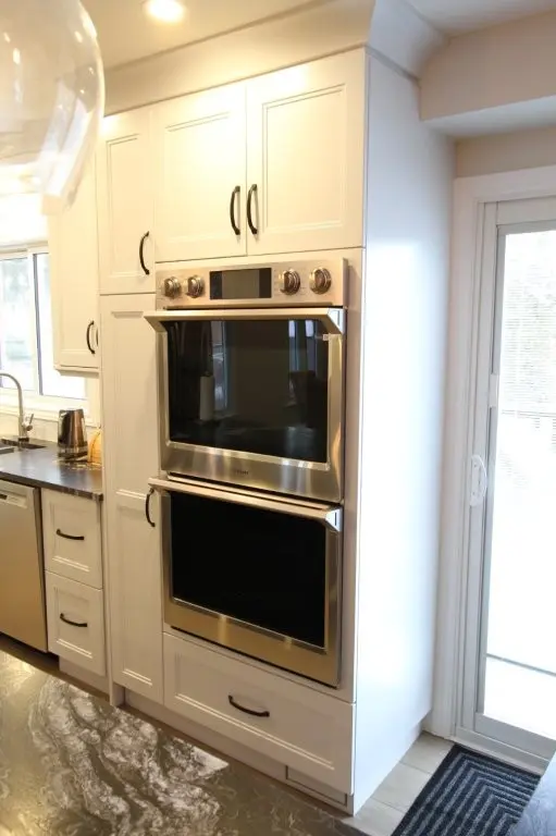 Modern kitchen with white cabinetry featuring built-in double stainless steel ovens, a dark countertop with a sink and faucet on the left, and a glass sliding door on the right with a striped black door mat.