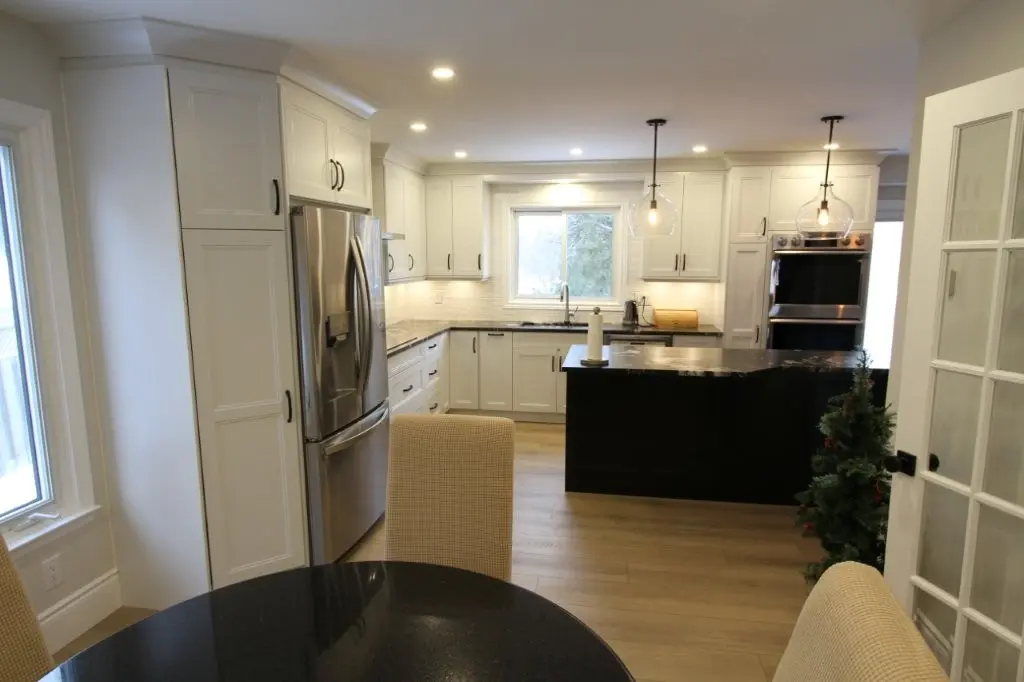 Modern kitchen with white cabinetry, a stainless steel refrigerator, black kitchen island with pendant lights above, and light wood flooring, seen from a dining area with beige upholstered chairs and a black round table.