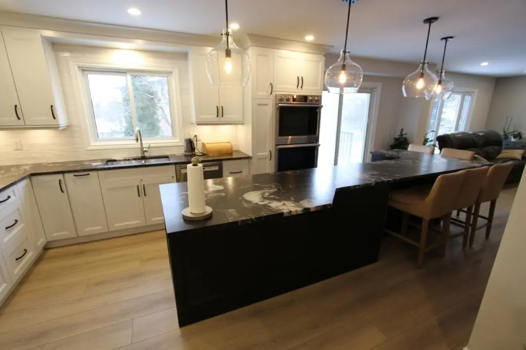 Modern kitchen with white cabinets, a large black granite island with a paper towel holder, three pendant lights hanging above the island, and wooden floor, with a view of a window and sliding glass doors in the background.