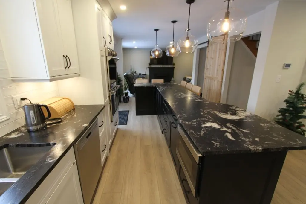 Modern kitchen interior with black granite countertops on white and black cabinets, stainless steel appliances, wooden flooring, pendant lights, and view into a living area with a fireplace.