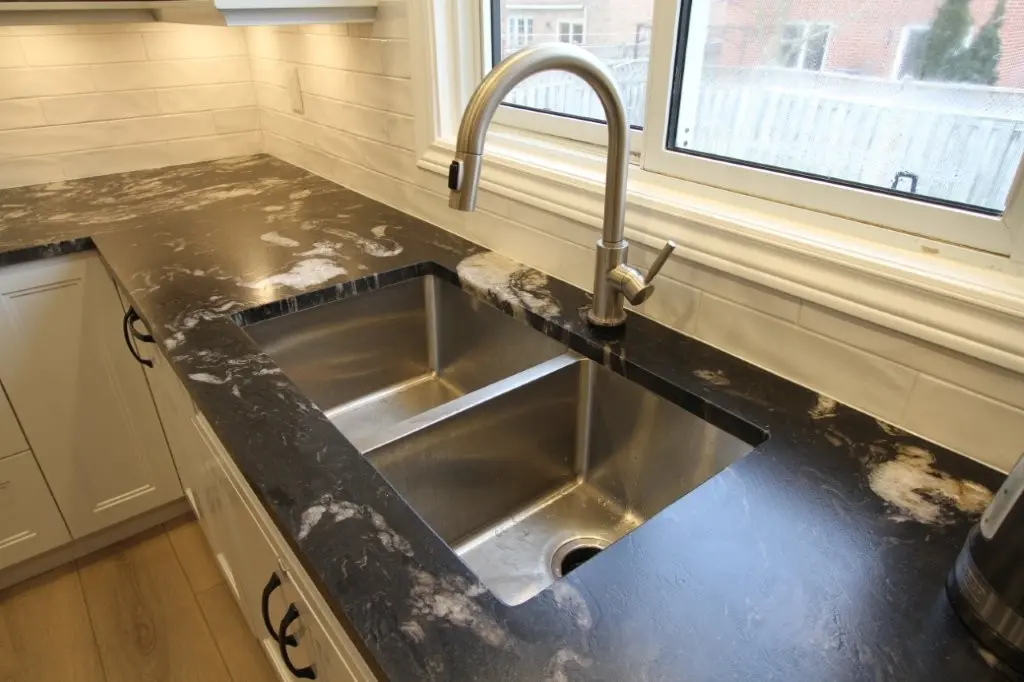 Modern kitchen sink with a brushed metal double-basin set into a black and white granite countertop under a window, surrounded by white tiled backsplash and cabinetry with black handles.