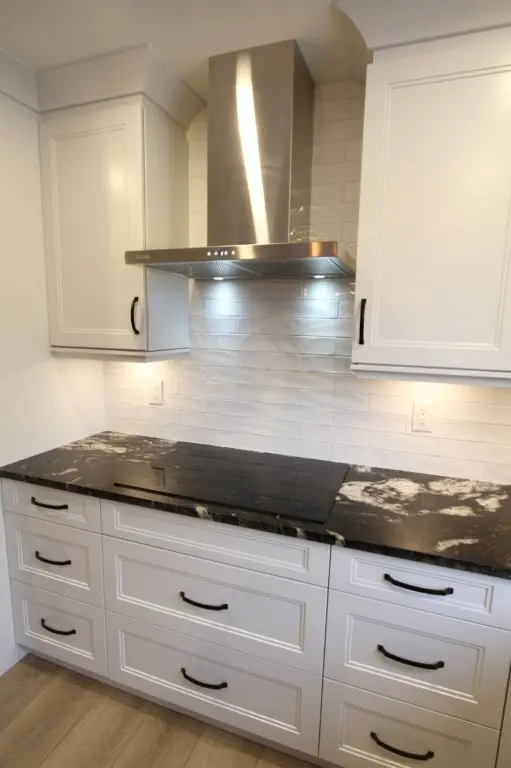Modern kitchen stovetop with black and white granite countertop, white cabinets with black handles, white subway tile backsplash, and stainless steel range hood with lights on.