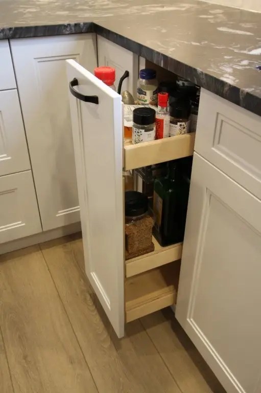 Open narrow kitchen cabinet drawer with multiple shelves containing jars and bottles of spices and condiments next to white cabinetry and a dark countertop with a wooden floor beneath.