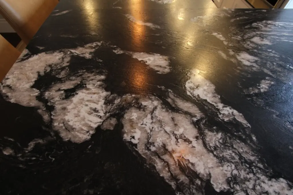 Close-up of a black granite surface with white and grey veining, reflecting ceiling lights, part of a kitchen or dining area with visible wooden chairs on one side.