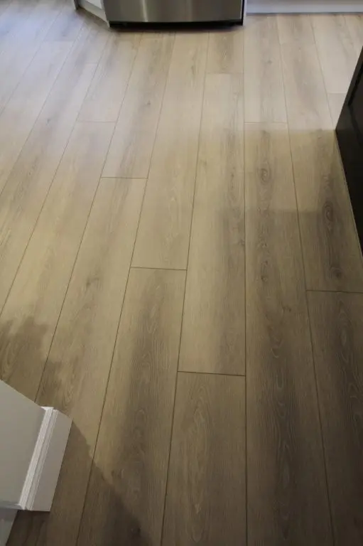 Light brown wooden laminate floor in a kitchen with part of a stainless steel appliance visible at the top and white and dark-colored cabinets on the sides.
