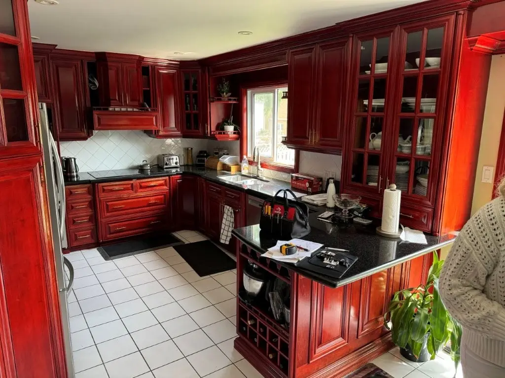 A before picture of a Kitchen with rich, dark red wooden cabinetry and black countertops, white tiled floor, appliances including a refrigerator, toaster, and kettle, window above a double sink, and various items on the counter including a black bag and papers.