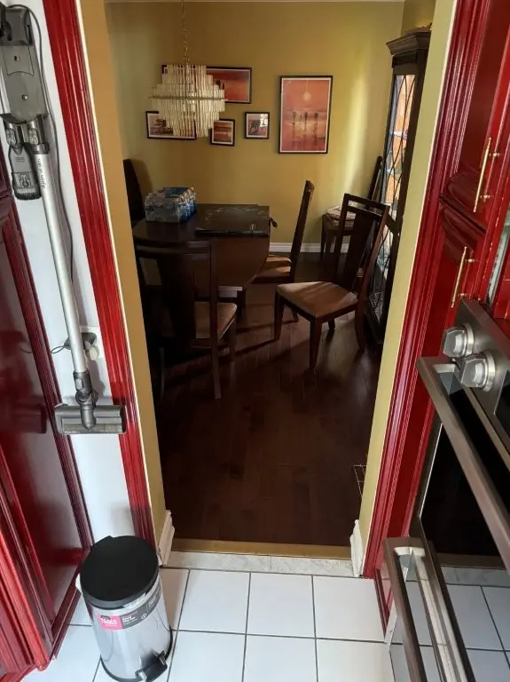 A before picture of a View through a doorway from a kitchen with red cabinets and stainless steel oven into a dining area with wooden floor, dark wooden table and chairs, a chandelier, and framed pictures on yellow walls.