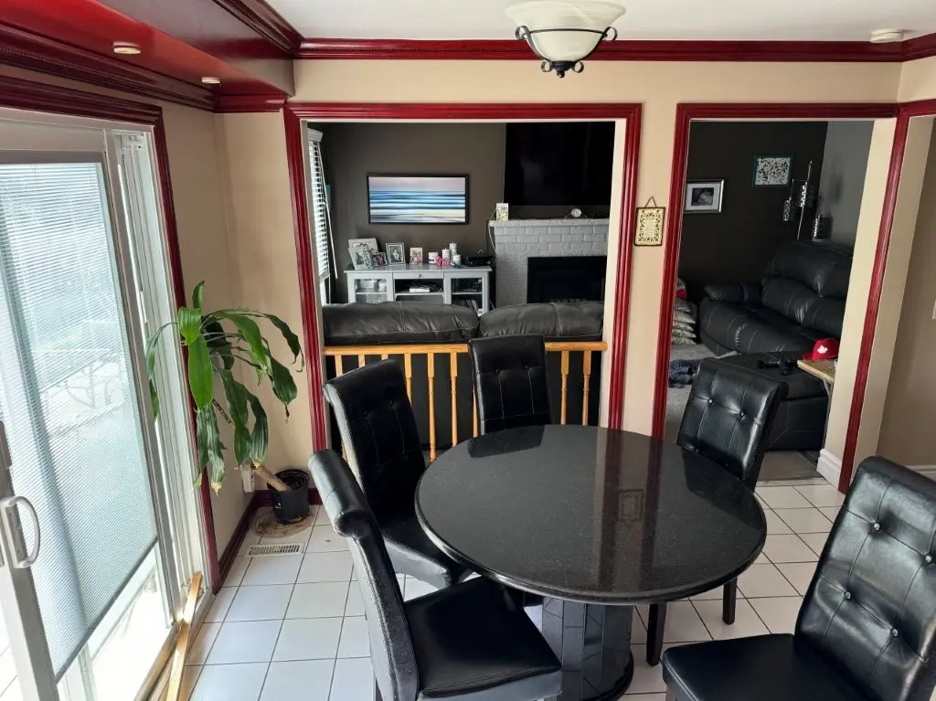 A before picture of a Dining area with a round black table surrounded by five black tufted chairs, adjacent to a sliding glass door with blinds and a potted plant, overlooking a living room with dark sofas, a white fireplace, and a flat-screen TV mounted above it.