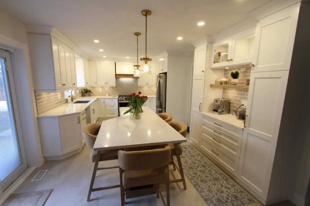 Modern kitchen with white cabinets, a long white island with beige chairs, pendant lights, and a vase of red tulips on the island counter, featuring a stainless steel refrigerator and a patterned rug along the floor on the right side.