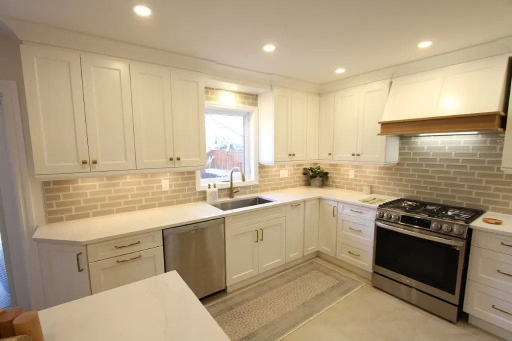 Bright modern kitchen with white cabinets, beige subway tile backsplash, stainless steel sink and dishwasher, stainless steel gas stove with a wooden-accented range hood, and a floor mat in front of the sink and stove area.