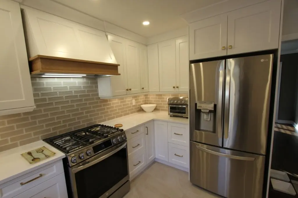 Modern kitchen corner with white cabinets, beige subway tile backsplash, stainless steel gas stove with range hood, stainless steel refrigerator, and a quartz countertop with a small oven and a bowl.