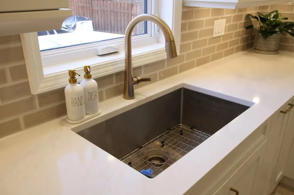 Modern kitchen sink with a large rectangular basin, bronze faucet, two white pump bottles labeled 'HANDS' and 'DISHES' on the left side, a tiled beige backsplash, and a small potted plant on the countertop to the right.