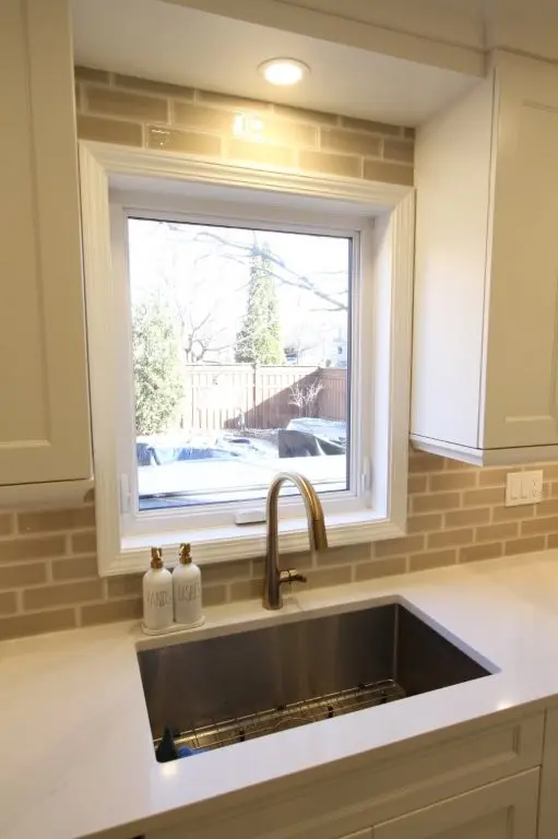 Modern kitchen sink area with a stainless steel undermount sink, brushed gold faucet, white countertop, light beige subway tile backsplash, and a window overlooking a backyard with a fence and trees
