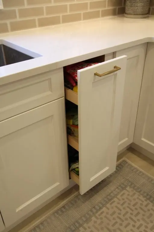 White kitchen cabinets with one narrow cabinet door slightly open revealing pull-out shelves with stored items, adjacent to a sink and beige tiled backsplash, with a patterned rug on the floor.