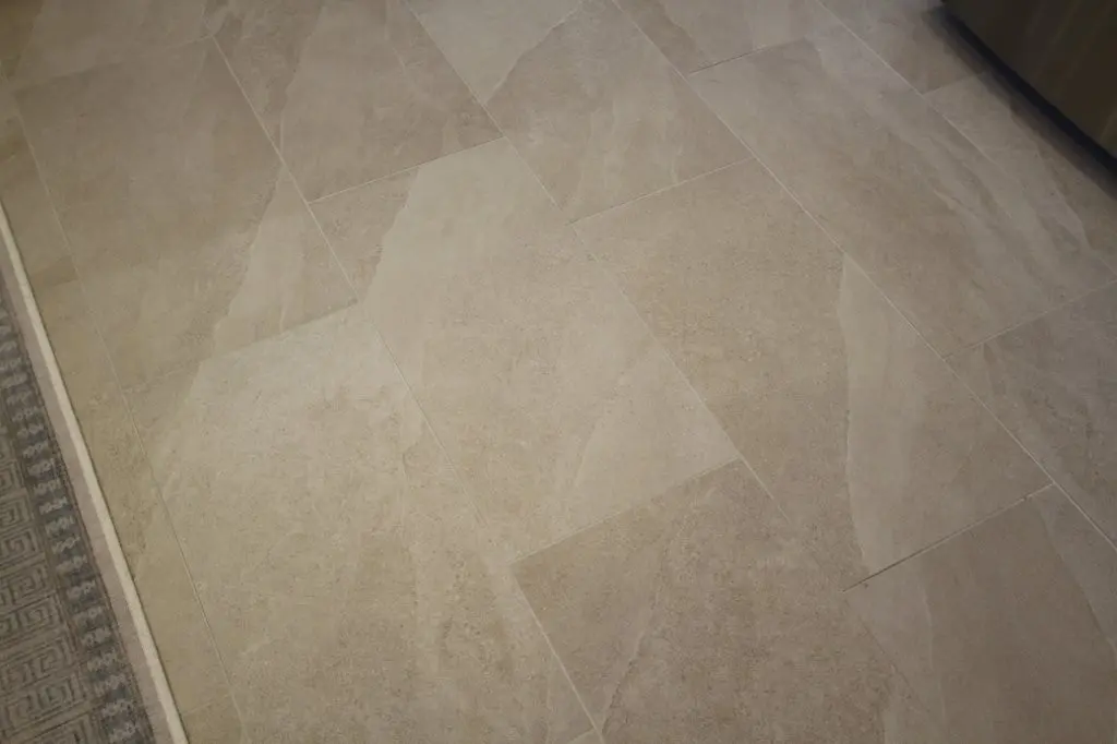 Close-up view of a beige tiled floor with a textured surface, showing the grout lines and part of a patterned carpet edge on the left side.