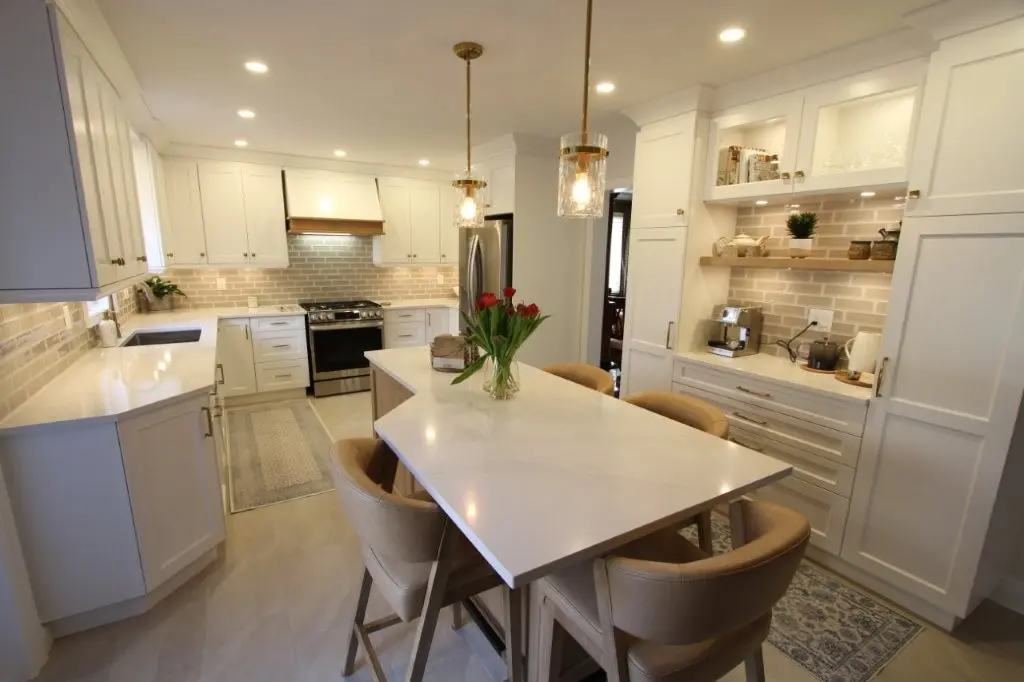 Modern kitchen with white cabinets, a large white quartz island with four tan cushioned chairs, pendant lights, stainless steel appliances, and a vase of red tulips on the island.