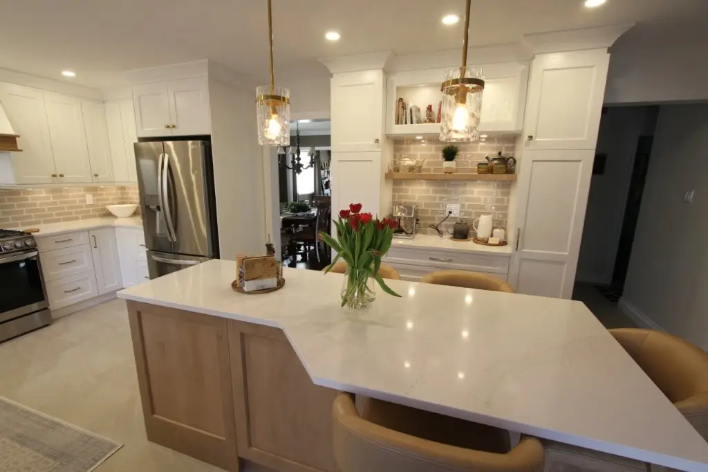 Modern kitchen with a white marble island countertop featuring a vase of red tulips, beige cushioned chairs, white cabinets, stainless steel refrigerator and stove, and a built-in shelf with kitchen accessories under warm pendant lighting.