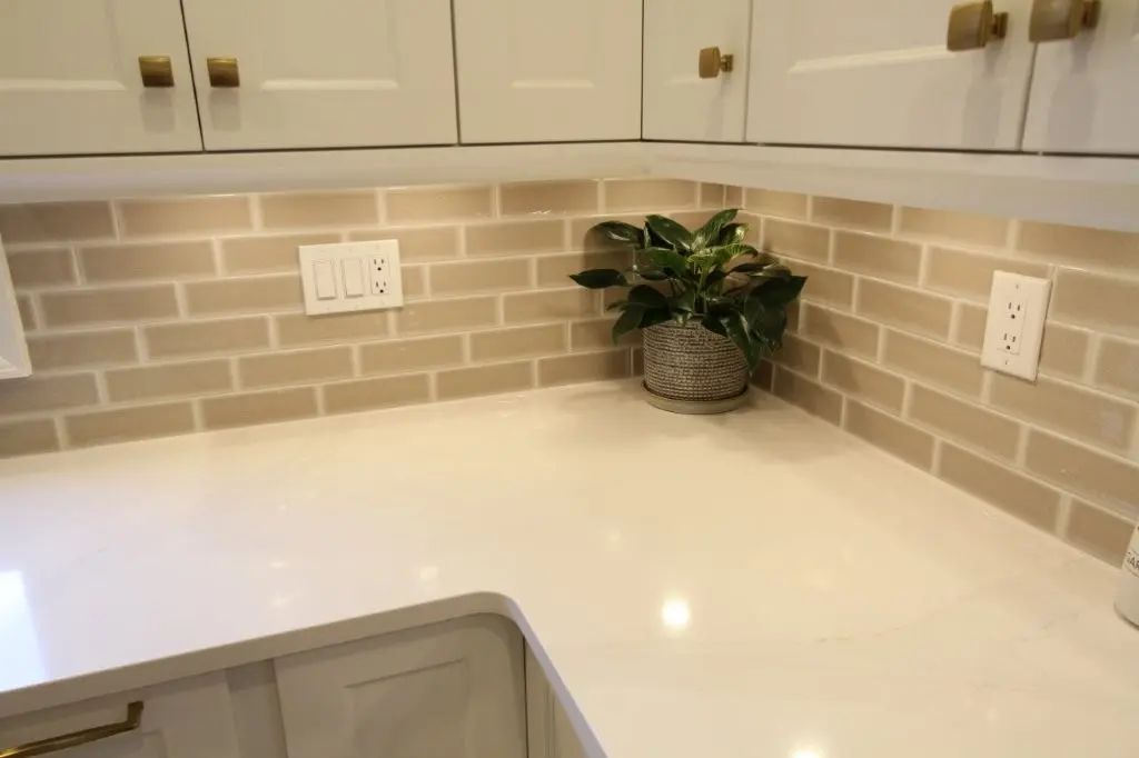 Corner of a kitchen countertop with a beige tile backsplash, white cabinets with brass handles, and a small green potted plant placed in the corner.