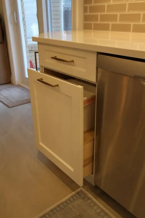 Partially open white kitchen cabinet drawer next to a stainless steel dishwasher, with beige tiled floor and brick-patterned backsplash in the background near a glass door.