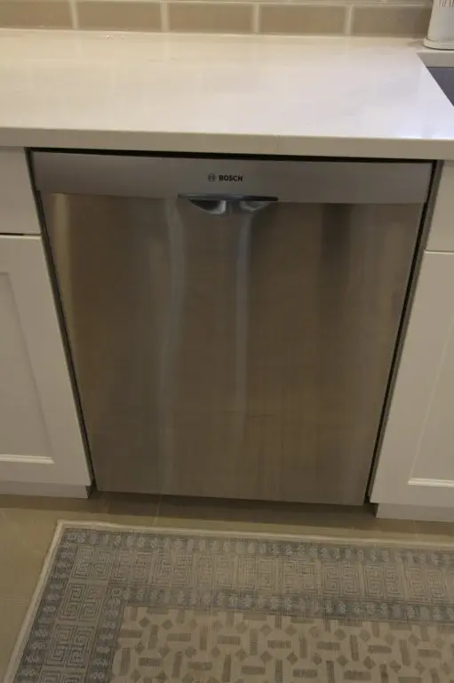 Stainless steel Bosch dishwasher integrated into white kitchen cabinetry under a white countertop with a beige tiled backsplash and a patterned rug on the floor in front.