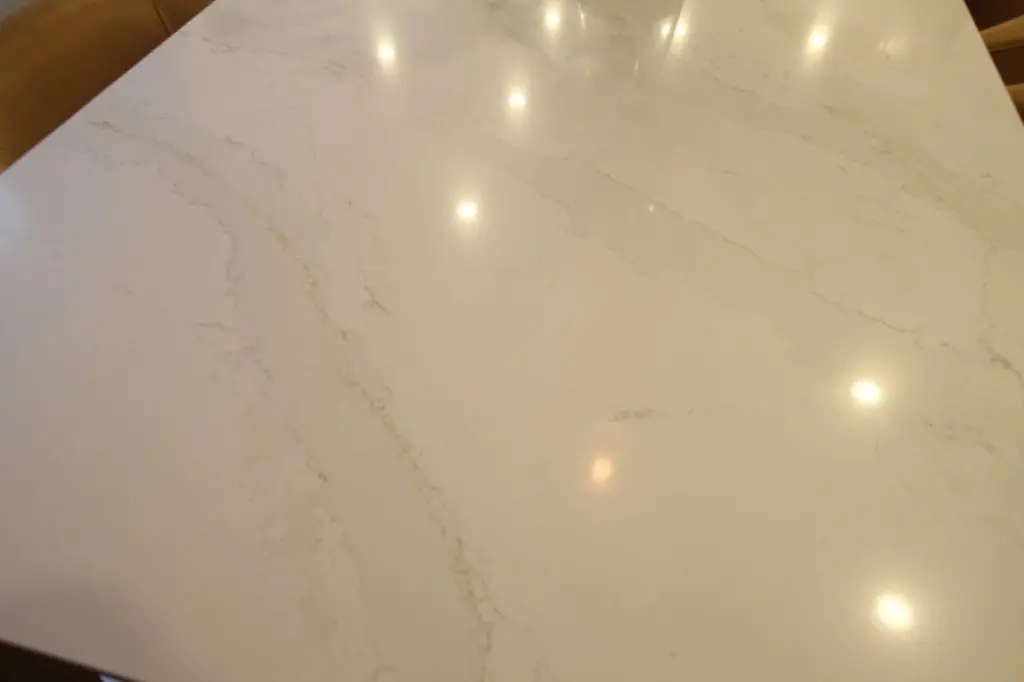 A smooth, polished white quartz table surface with subtle light grey and beige veining, reflecting several ceiling lights.