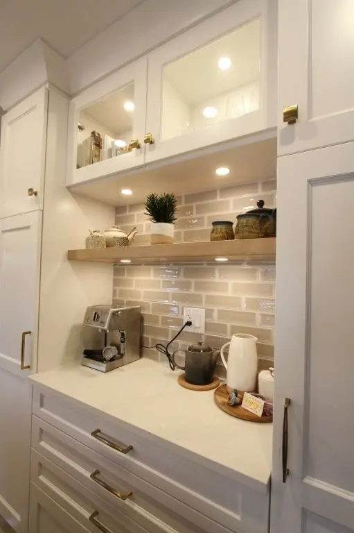 A modern kitchen coffee nook with white cabinetry, a light wood floating shelf holding jars and a small plant, a silver espresso machine on a white marble countertop, a gray mug on a coaster, and a wooden tray with a white pitcher, sugar bowl, and coffee pods against a beige subway tile backsplash