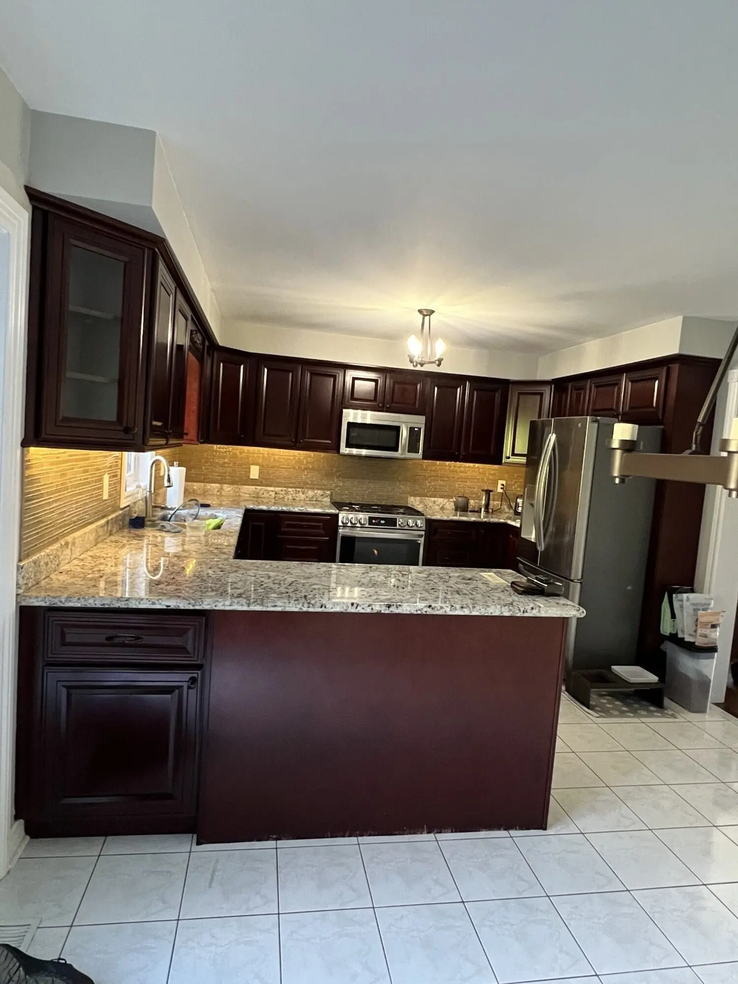Before picture of a kitchen with dark wood cabinets, granite countertops, tiled floor, stainless steel appliances including a refrigerator, microwave, and stove, and under-cabinet lighting along the backsplash with a central ceiling light fixture.