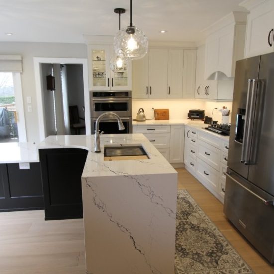 Modern kitchen with white quartz countertops, black lower cabinets, white upper cabinets, stainless steel refrigerator, built-in ovens, gas stove, and two pendant lights over the island with a sink, light wood flooring, and a patterned rug near the stove.