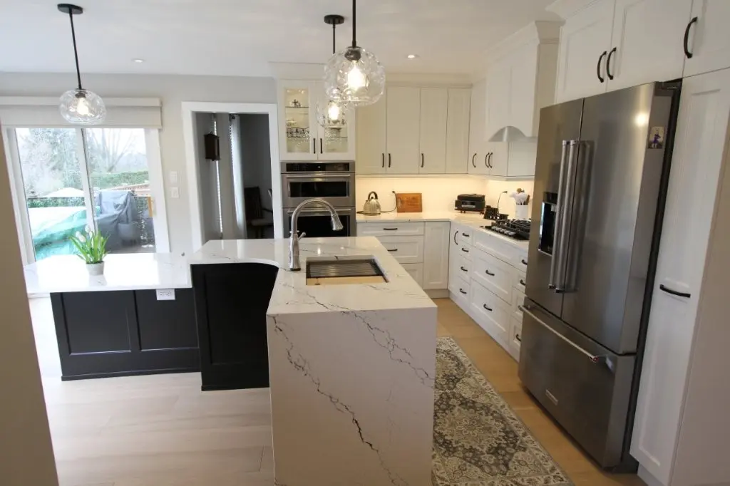 Modern kitchen with white quartz countertops, black lower cabinets, white upper cabinets, stainless steel refrigerator, built-in ovens, gas stove, and two pendant lights over the island with a sink, light wood flooring, and a patterned rug near the stove.