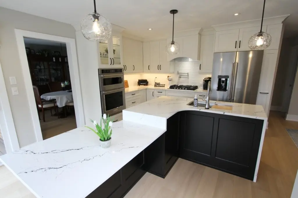 Modern kitchen with white quartz countertops and black lower cabinets, pendant lights hanging over the island, stainless steel double oven and refrigerator, and an adjacent dining room with a round table and chairs.