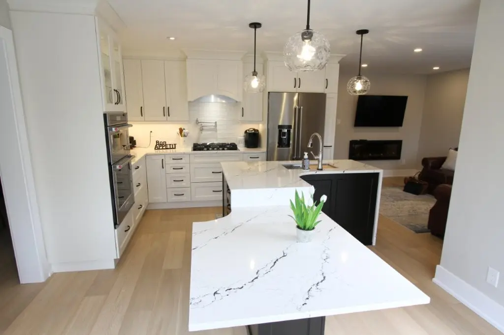 Modern kitchen with white cabinets, a quartz-topped island with a small potted plant, stainless steel appliances, three round pendant lights, light wood flooring, and a living room area visible in the background with a mounted TV and fireplace.