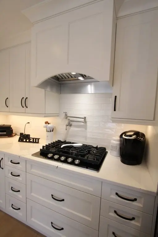 White modern kitchen with a gas stovetop, white backsplash tiles, ivory countertops, black handles on cabinets and drawers, a coffee maker, container labeled 'COFFEE', and overhead range hood with built-in lighting.