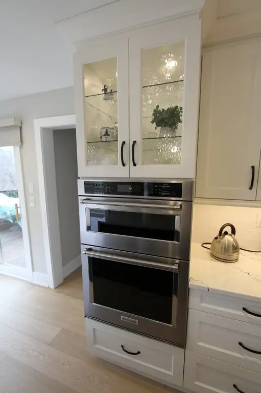 Built-in stainless steel double oven with digital controls in white kitchen cabinetry, next to a quartz countertop with a stainless steel kettle and glass-front cabinets above with decorative items inside.