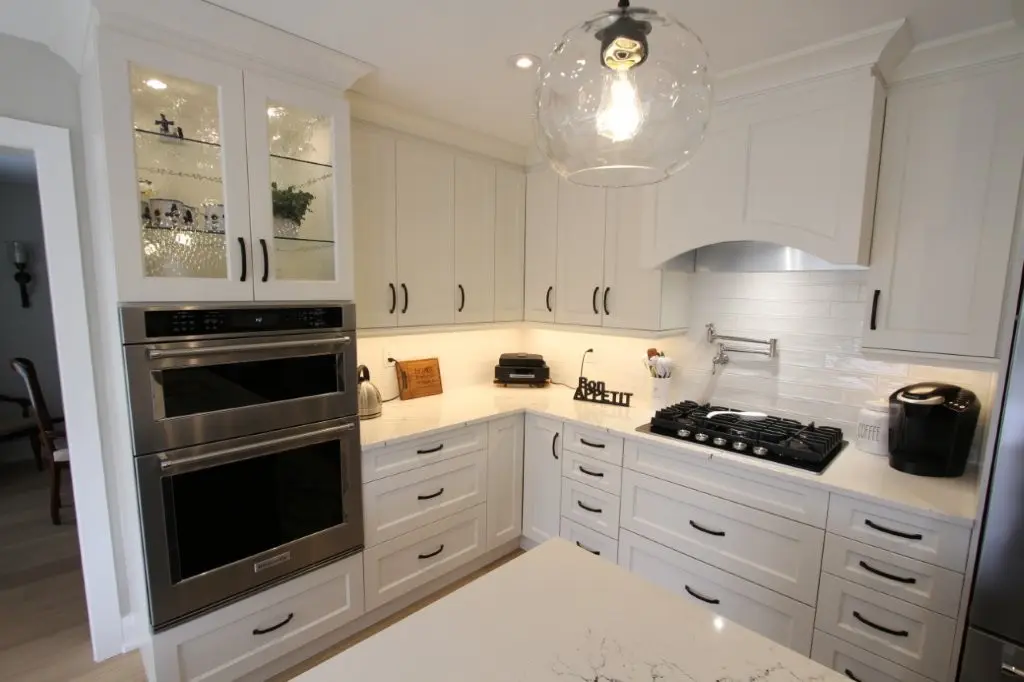 Modern white kitchen with built-in double oven, gas stove with hood, white cabinets with black handles, illuminated glass-front cabinet, coffee maker, and a large clear glass pendant light hanging from the ceiling