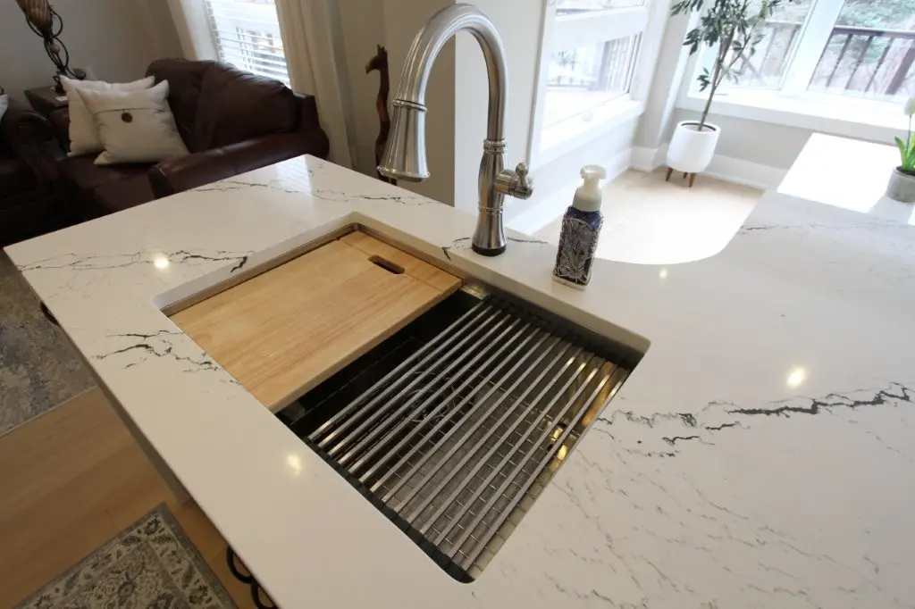 Modern kitchen sink with a stainless steel faucet, a wooden cutting board partially covering the sink, and a soap dispenser on a white quartz countertop with black veins, adjacent to a living room with brown leather couches and large windows.