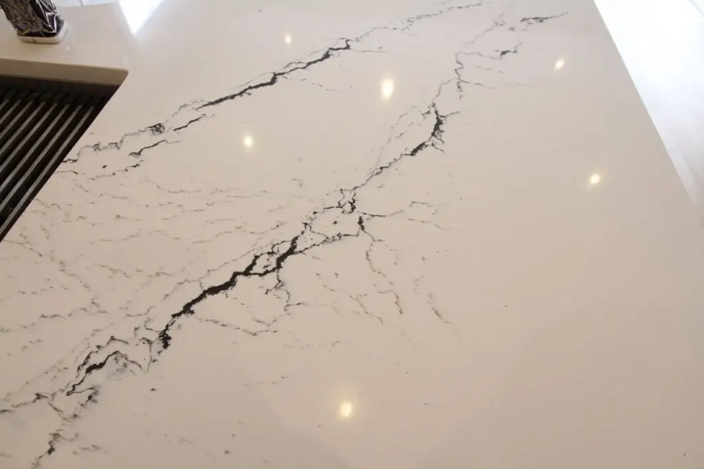 Close-up of a white quartz countertop with black veining and reflections of ceiling lights, alongside a portion of a kitchen sink area.