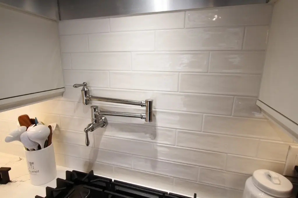 Kitchen backsplash with white rectangular tiles, a chrome pot filler faucet mounted on the wall above a gas stove, a white container with cooking utensils on the left side, and a white ceramic canister on the right side.