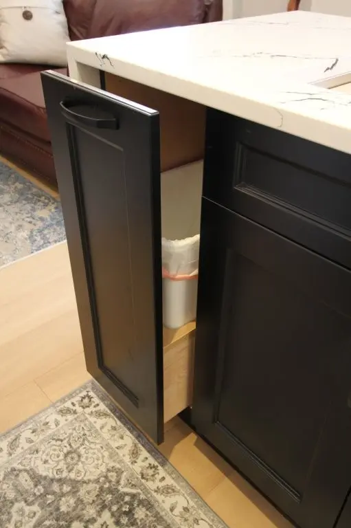 Black kitchen cabinet door partially open revealing a pull-out trash bin with a lined white trash can inside, set against a light wood floor and a quartz countertop.