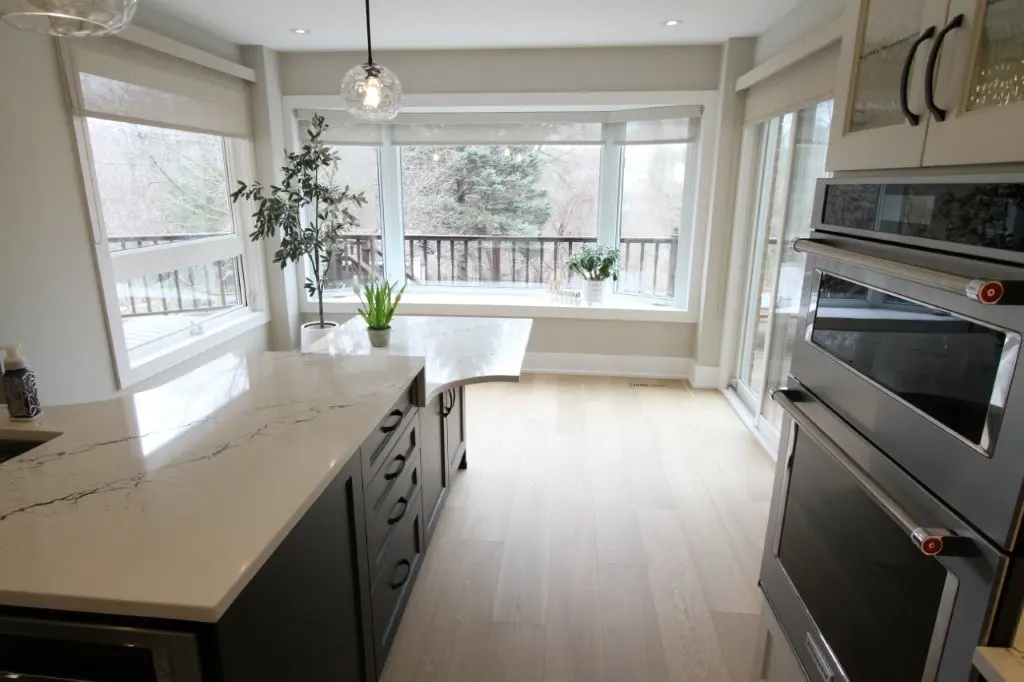 Bright modern kitchen corner with light wood flooring, a quartz countertop island with drawers, large bay windows with a view of trees, a potted plant on the countertop, and stainless steel built-in ovens.