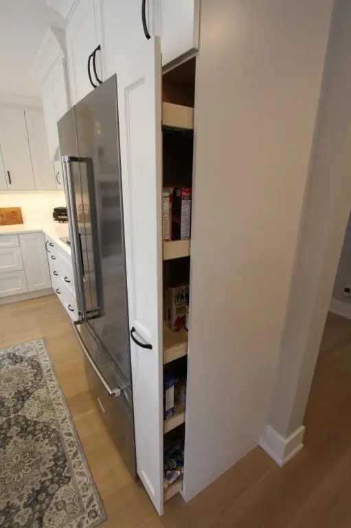 Tall, narrow kitchen pantry cabinet door open, showing shelves stocked with cereal boxes and other food items beside a stainless steel refrigerator in a white kitchen with light wood flooring and a patterned rug.