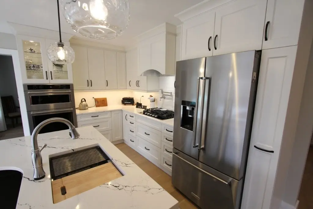 Modern kitchen with white cabinetry, stainless steel refrigerator, built-in oven, gas stove, quartz countertop island with a sink and cutting board, and two pendant lights hanging from the ceiling.