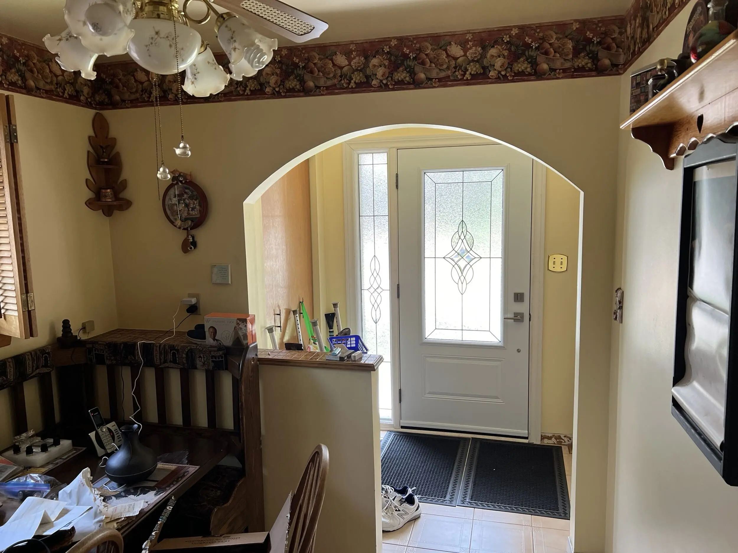 A before picture of an Entryway with a white door featuring decorative frosted glass panels, two black floor mats, and a pair of white sneakers on beige tile flooring, viewed through a beige-painted archway with a wooden bench and cluttered table on the left side and a wall-mounted wooden shelf on the right side, under a ceiling fan with floral-patterned glass light shades and fruit-themed wallpaper border near the ceiling.