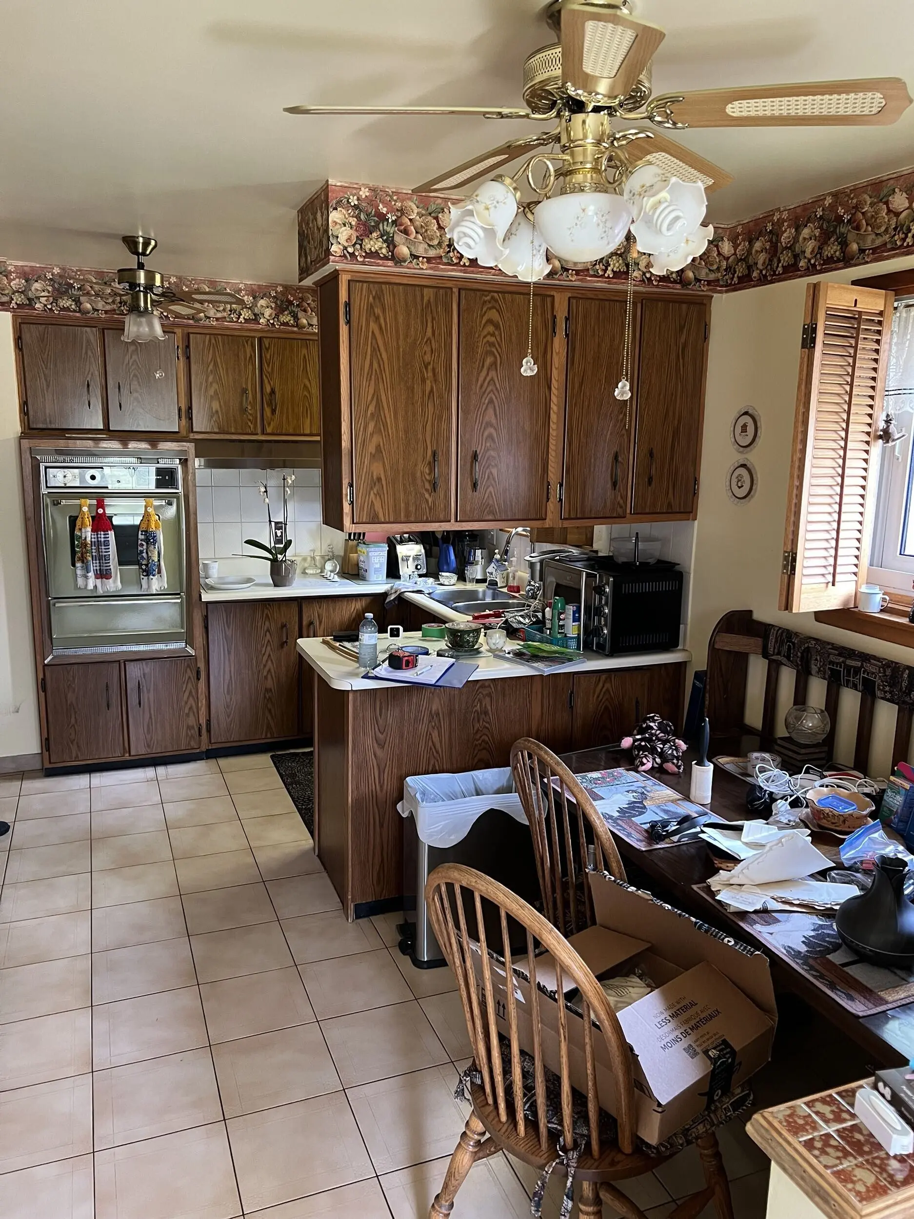 before photo of a Retro-style kitchen with wood-paneled cabinets, floral wallpaper border, and tiled floor, featuring a ceiling fan with floral light fixtures, a countertop cluttered with various items, a dining table with chairs, and an open window with wooden shutters.